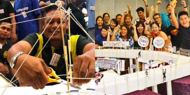 DBD HUB corporate wellness team building activity in Singapore showing employees collaborating in Bridging Teamwork using satay sticks to construct a strong, stable bridge—focusing on leadership, teamwork, SCRUM principles, project management, and communication under time pressure.